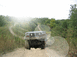 1986 Ford Bronco - An SUV travels down a dusty, unpaved road surrounded by dense foliage under a bright sky.