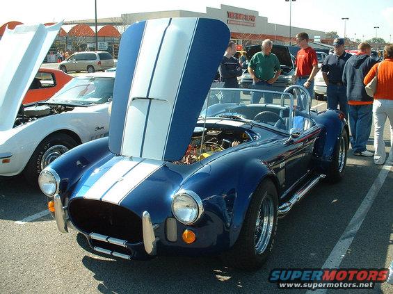 Blue AC Cobra replica with hood open, white stripes on hood and body. White Corvette with hood open to the left. People standing around
