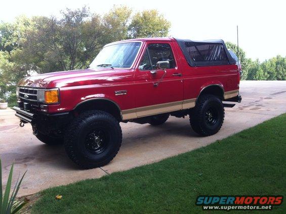 Red 1987 Ford Bronco with tan side trim, black soft top, and lifted suspension parked on a concrete driveway