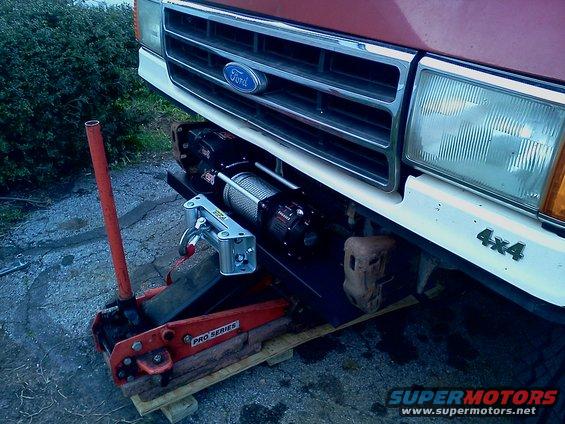 Ford truck front end, grille, headlights, and a winch mounted below the bumper. A red floor jack is positioned in front of the winch