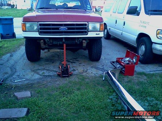 Red Ford truck with a jack under the front, parked next to a white van. A bumper lies on the grass