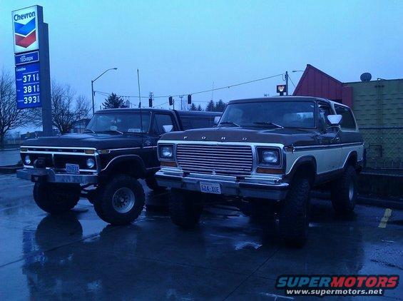 Two lifted Ford Broncos parked at a gas station, one brown and white, the other black