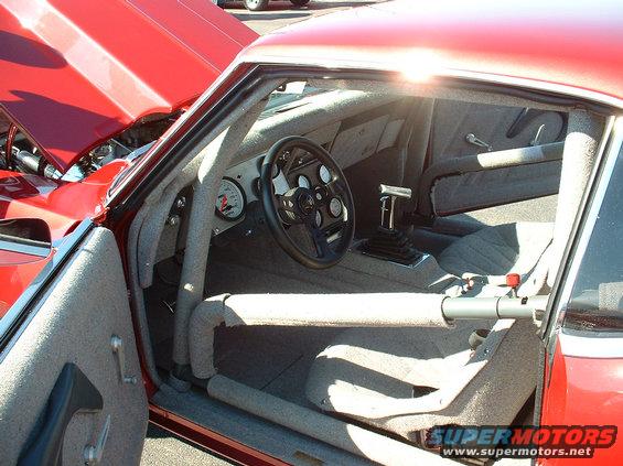 Interior of a red race car with roll cage, racing seats, and aftermarket steering wheel. Sun glare is visible