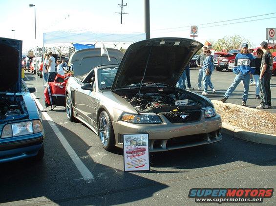 Tan Ford Mustang convertible with hood open, parked next to a blue car. Sign in front says '2001 Select Masters'
