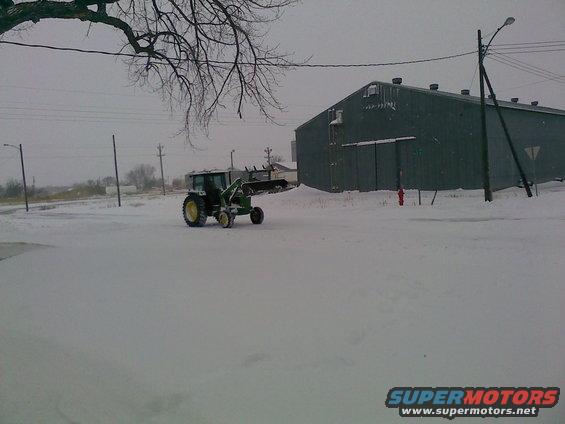 Green tractor with front loader in snow, next to a large building