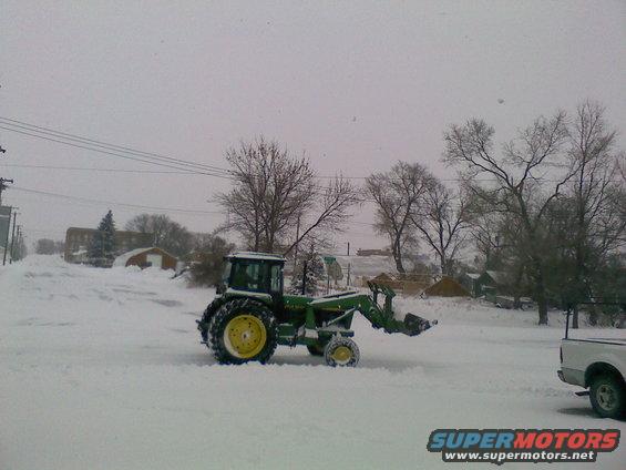 Green tractor with front loader in snow, partial view of white pickup truck