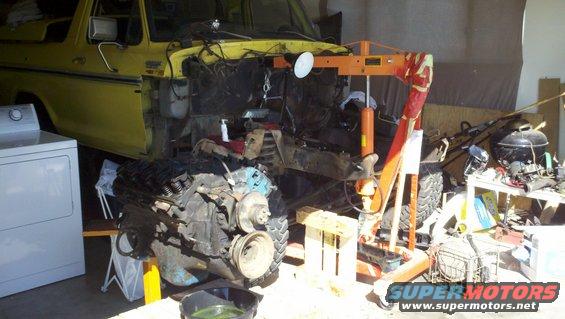 Yellow 1978 Ford Bronco with engine removed by an orange engine hoist, parked next to a white washing machine