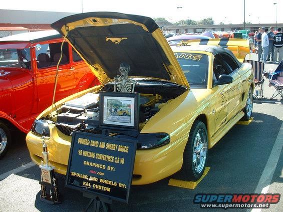 Yellow 1998 Ford Mustang convertible with hood open, displaying a skeleton and a sign detailing owners and modifications