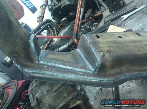 Welded metal frame section with engine components and exhaust pipes in background