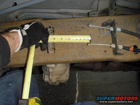 Gloved hand holding tape measure against rusty vehicle frame, measuring a section near brake cables