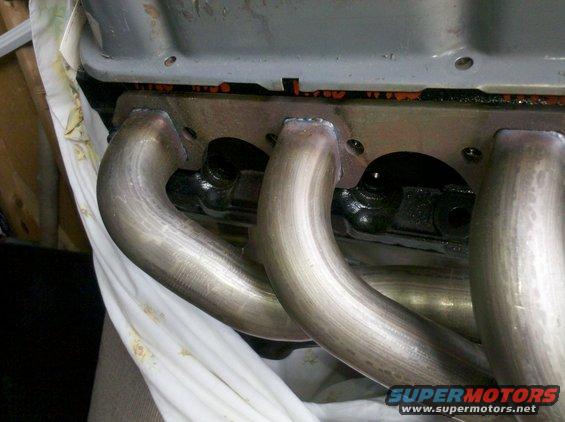 Exhaust manifold pipes connected to an engine block, with orange gasket material visible