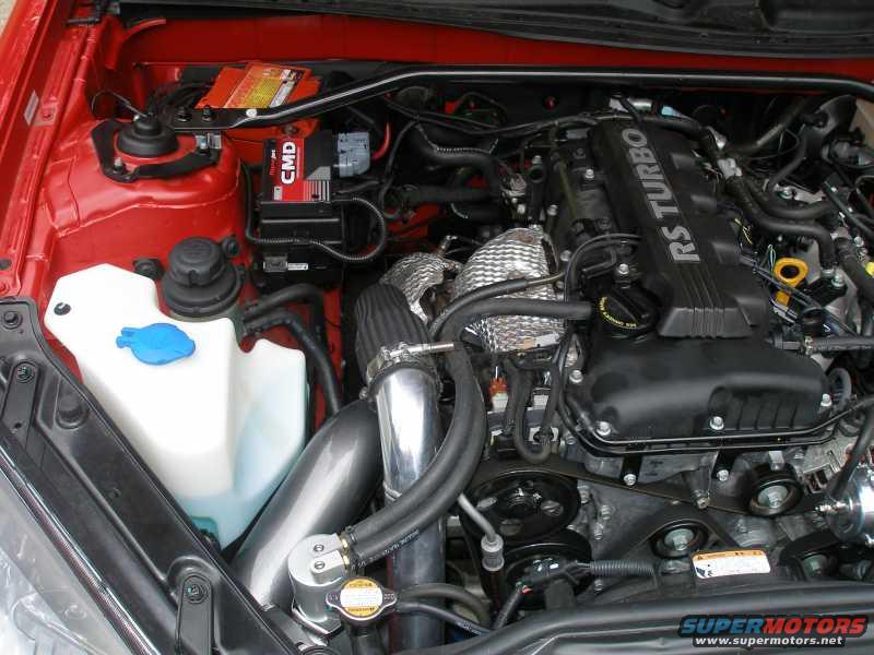 Engine bay with a black engine cover labeled "RS TURBO", a white fluid reservoir, and a red battery
