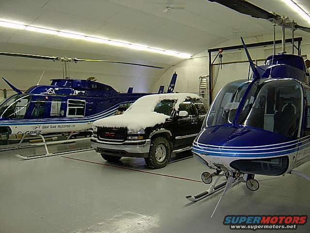 Black 1999 GMC Sierra 1500 pickup truck covered in snow, parked in a hangar with helicopters