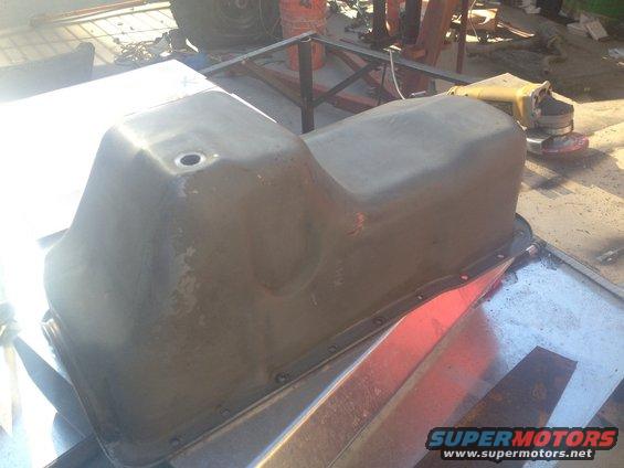 Gray metal oil pan with a hole on top, resting on a metal surface. A grinder is visible in the background