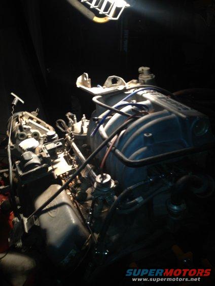 Engine components under a work light, with blue and black hoses. Some text visible on the engine block