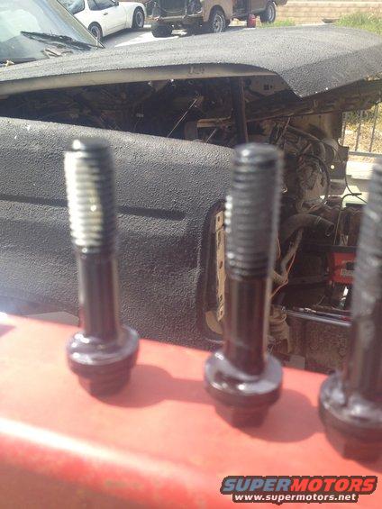 Three black bolts in focus, with an engine bay and other vehicles blurred in the background
