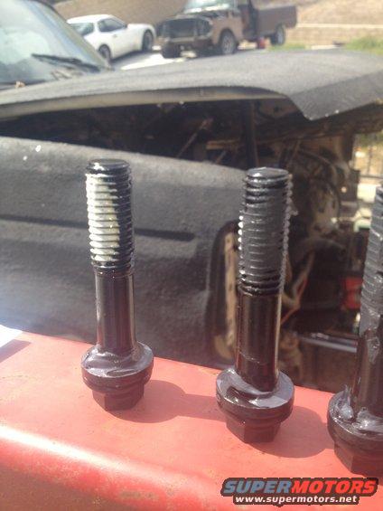 Close-up of bolts with white sealant on a red surface, with a vehicle engine and other cars in the background