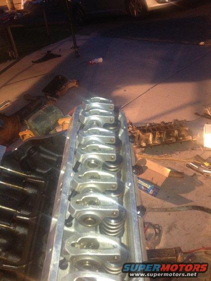 Close-up of an engine cylinder head with rocker arms and springs visible. Other engine parts and tools are scattered on the ground in the background