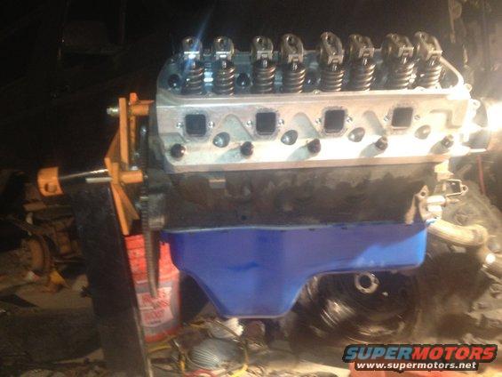 Engine block with blue oil pan and silver cylinder heads with valve train visible, mounted on an engine stand