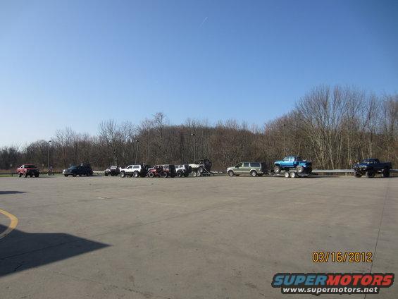 Several off-road vehicles and trucks parked in a lot, some on trailers, under a clear blue sky