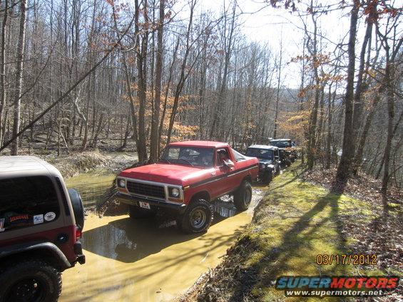 Red 1979 Ford Bronco driving through a muddy trail with other off-road vehicles behind it