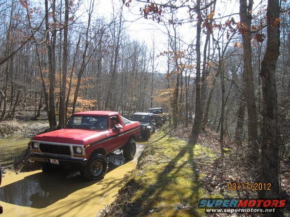 Red 1979 Ford Bronco driving through a muddy creek bed with trees on either side