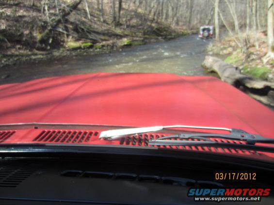 View from inside a red vehicle looking out at another SUV crossing a creek in a wooded area