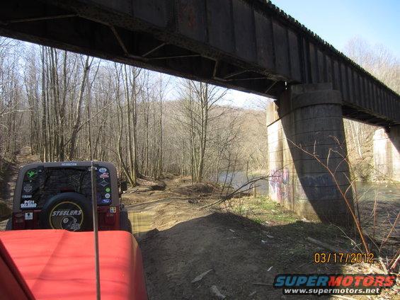Two Jeeps on a muddy trail under a large railway bridge, trees and a river in background