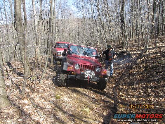 Red Jeep Wrangler and red Ford pickup truck on a dirt path in a wooded area