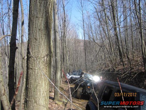 Several off-road vehicles driving on a narrow dirt trail through a dense forest. Date stamp reads 03/17/2012
