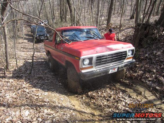 Red SUV driving on a muddy trail in woods. Another SUV is behind it