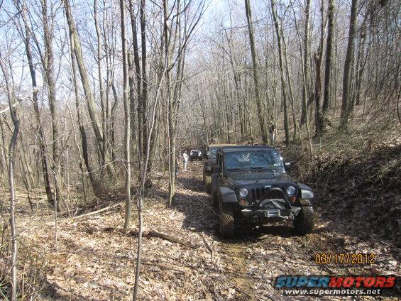 Black Jeep Wrangler on a muddy forest trail, with other vehicles behind