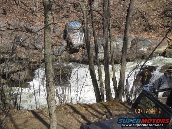 Black vehicle's front corner and windshield are visible, parked on a dirt path next to a waterfall
