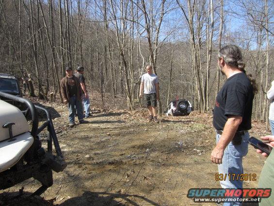 Several people stand around off-road vehicles in a wooded area; one vehicle is overturned