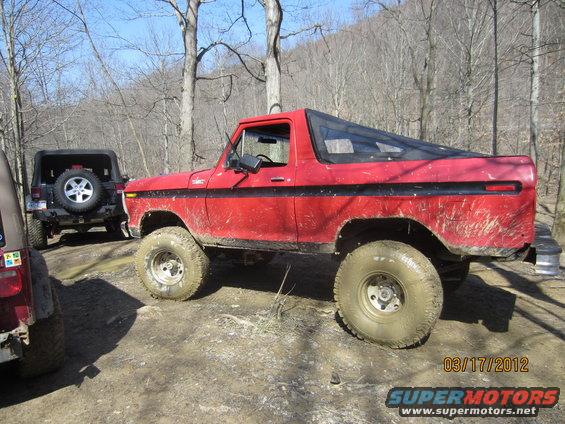 Red 1978 Ford Bronco with black trim and a black soft top, seen from the side on a dirt road