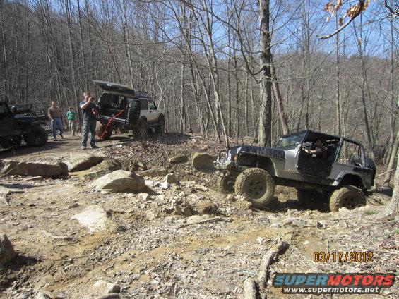 Several Jeeps navigating a rocky, wooded trail. One Jeep is in the foreground, angled to the right