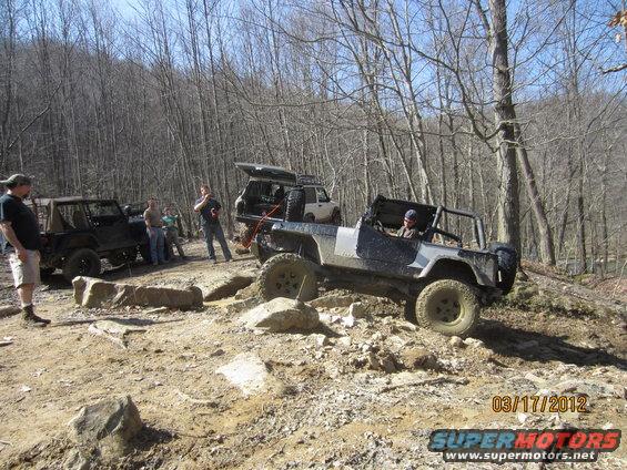 Several off-road vehicles are navigating a rocky, wooded trail. One Jeep is in the foreground, going over rocks