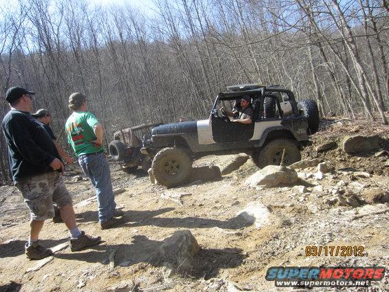 Two off-road Jeeps navigating a rocky trail with people watching. One Jeep is in the foreground, the other behind it