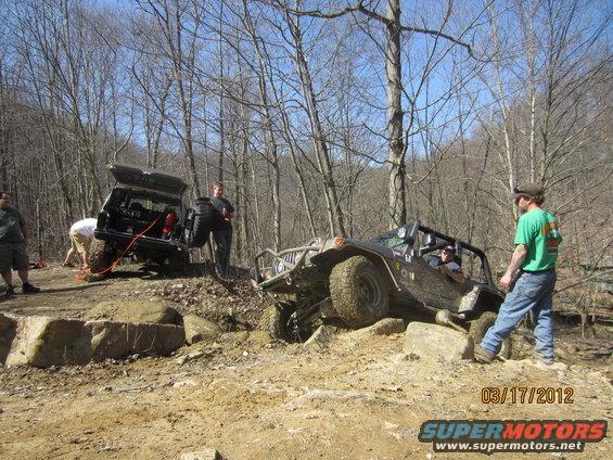 Two off-road vehicles are on a dirt trail with trees around. One vehicle is on its side, and people are working on it. The other vehicle is in the foreground, also on a rocky path