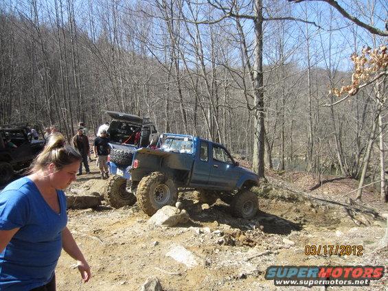 Several modified off-road trucks navigating a rocky, wooded trail. One blue truck is prominently featured on the right