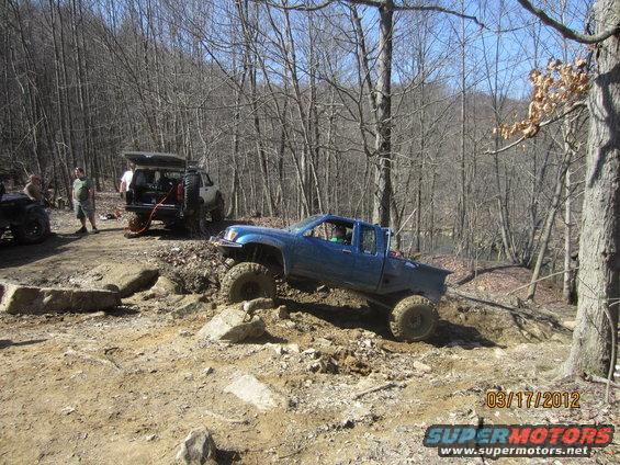 Blue pickup truck with large tires navigating a rocky trail. Another SUV is parked nearby with its trunk open