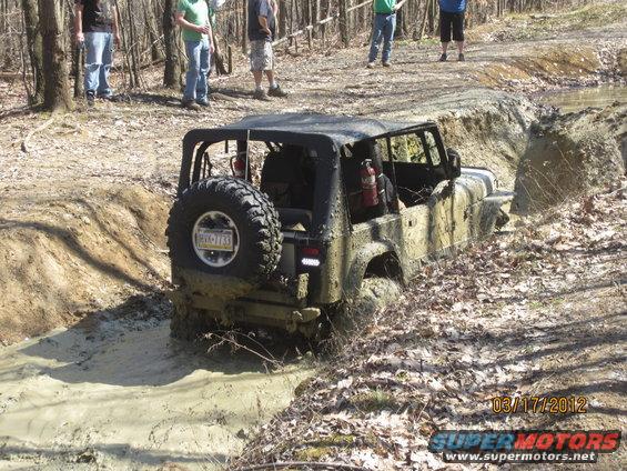 Mud-splattered Jeep Wrangler with spare tire on rear, stuck in a muddy ditch