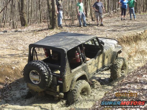Muddy Jeep Wrangler with soft top stuck in a muddy ditch, people watching from the bank