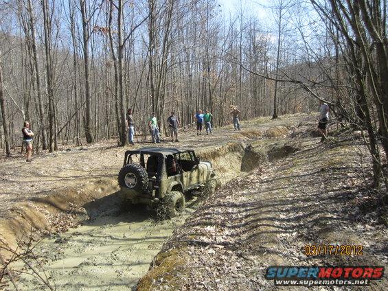 Mud-splattered Jeep Wrangler navigating a muddy ditch in a wooded area