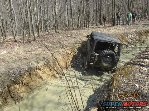 Jeep Wrangler driving through a muddy trail in a forest. People watch from the side
