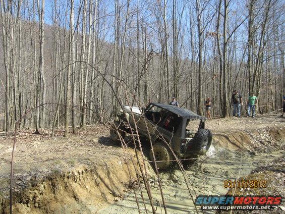 Modified off-road vehicle stuck in a muddy trail with people watching