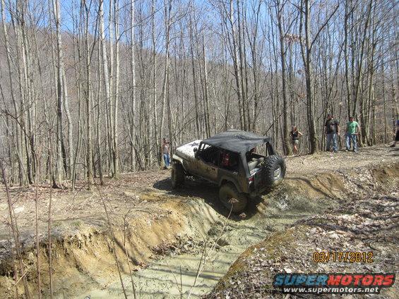 Tan Jeep Wrangler with a black soft top navigating a muddy trail, rear-mounted spare tire visible
