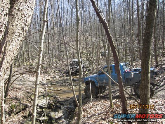 Blue pickup truck and another off-road vehicle driving through a wooded area with a stream