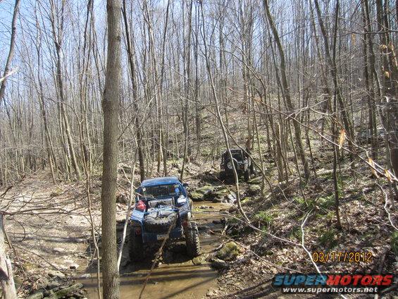 Two off-road vehicles navigating a rocky stream bed in a wooded area