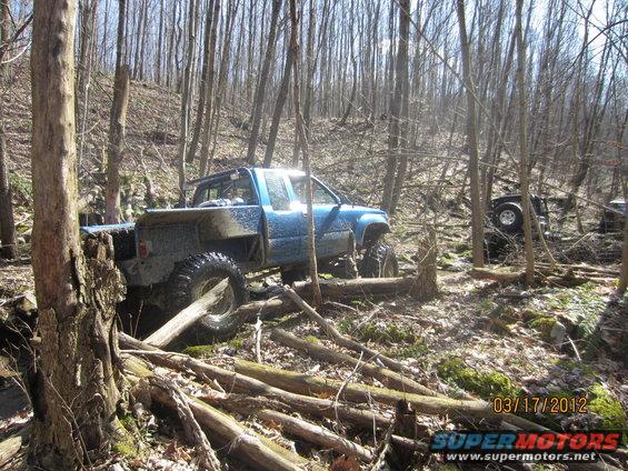 Blue pickup truck with large tires off-roading in a forest, with another vehicle visible in the background